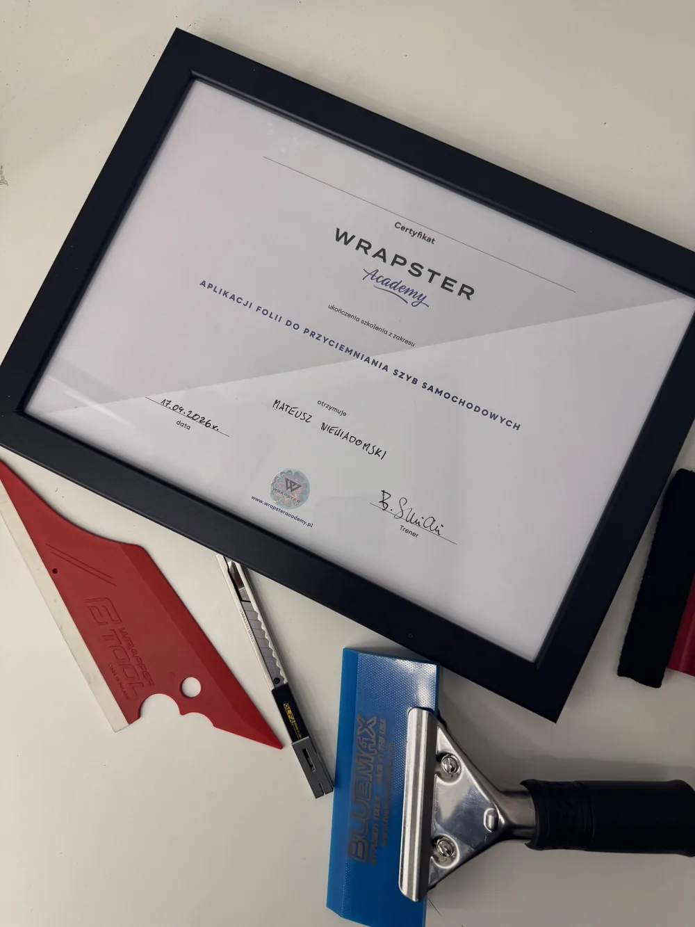 Certyfikat Wrapster Academy ukończenia szkolenia z zakresu aplikacji folii do przyciemniania szyb samochodowych, data 17.04.2026, w ramce obok narzędzi do montażu folii